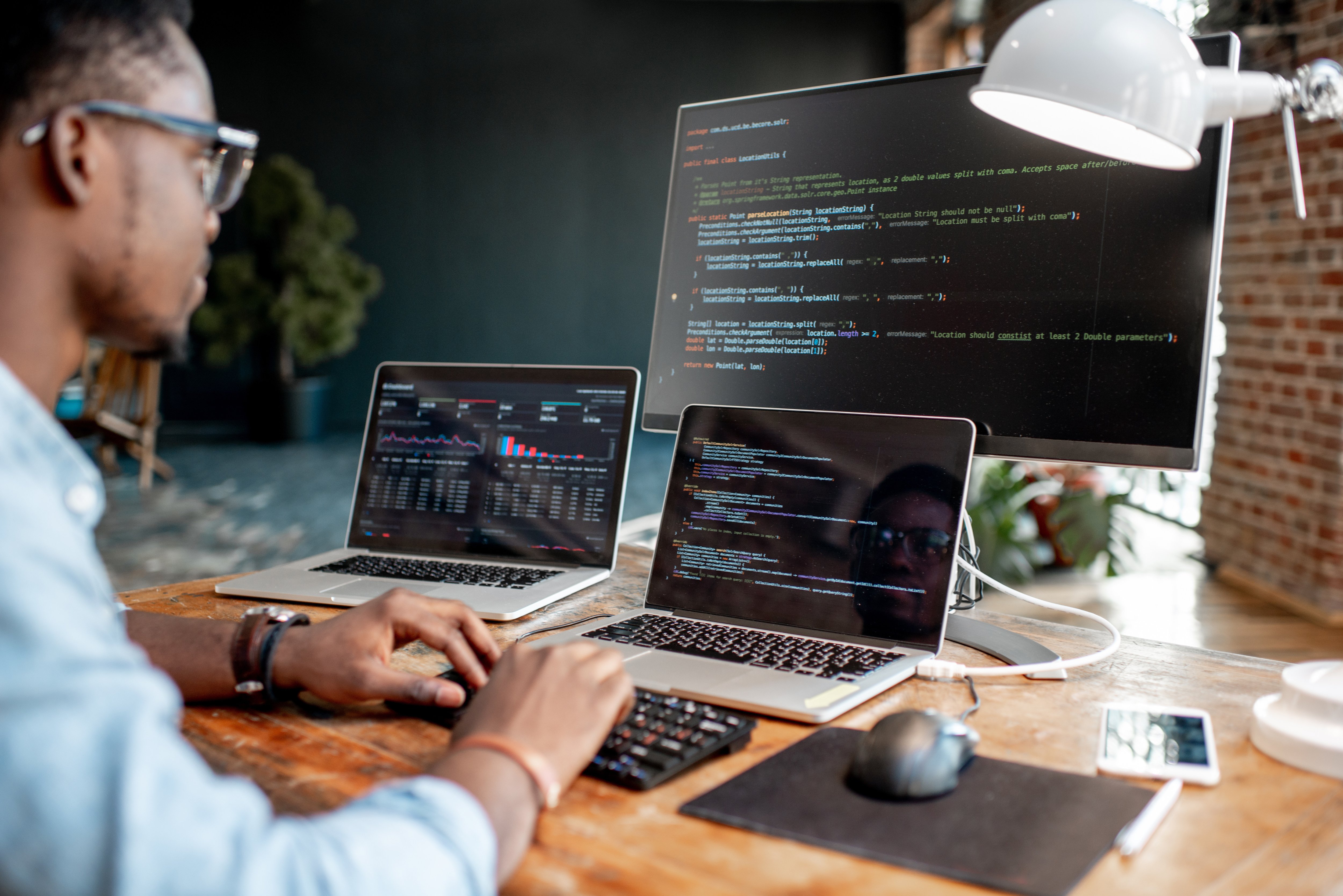 Young male programmer working at a wooden desk, typing on a black keyboard, facing two laptops and a large monitor